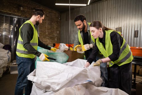 Multiethnic workers in safety vests and protective gloves separating garbage near sacks while